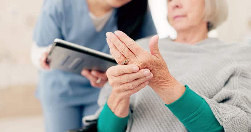 A Close-Up On The Hands Of A Senior Woman, Who Is Rubbing Her Knuckles Due To Joint Pain From Arthritis. In The Background, A Female Doctor Holds A Tablet During A Consultation.