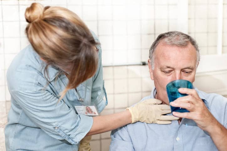 A Female Speech Therapist, Wearing Gloves, Places Her Hand On A Senior Man'S Throat To Monitor His Swallowing As He Drinks From A Blue Cup During A Dysphagia Assessment.