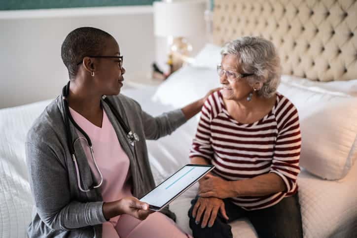 A Female Doctor With A Stethoscope Holds A Tablet And Speaks Compassionately To A Senior Woman. They Are Sitting Together On A Bed, And The Doctor Has A Reassuring Hand On The Patient'S Shoulder During An In-Home Visit