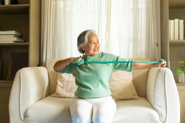 A Smiling Senior Asian Woman With Gray Hair Sits On A Sofa In Her Living Room, Performing An Arm Exercise By Stretching A Green Resistance Band.
