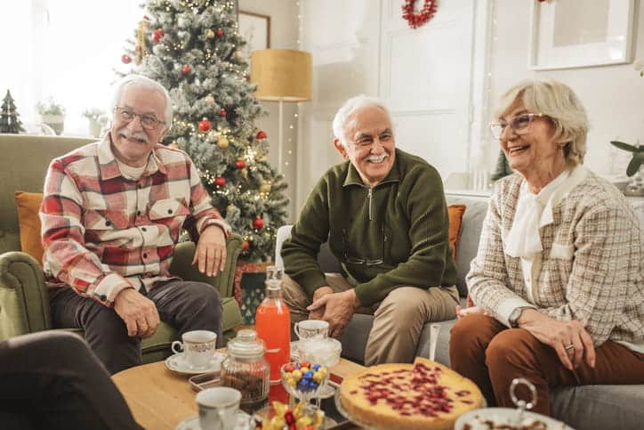 A Group Of Three Happy Seniors—Two Men And One Woman—Laugh While Sitting On A Couch And Armchair In A Living Room Decorated With A Christmas Tree.
