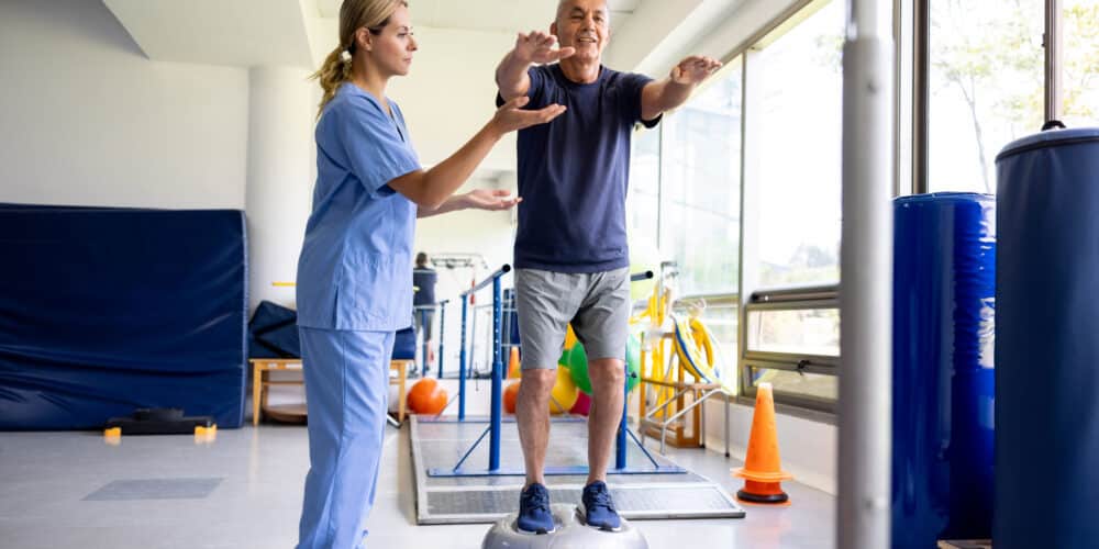 Mature Latin American Man Doing Exercises Using A Balance Ball With The Help Of His Physical Therapist