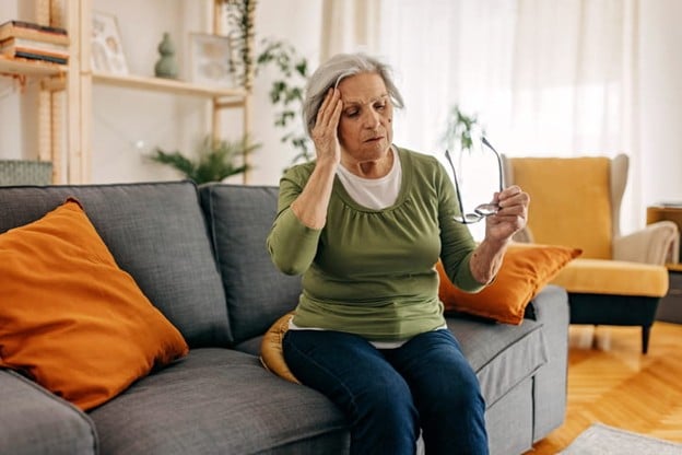 An Elderly Woman Holds Her Head As She Sits On The Couch Because She Is Dizzy.