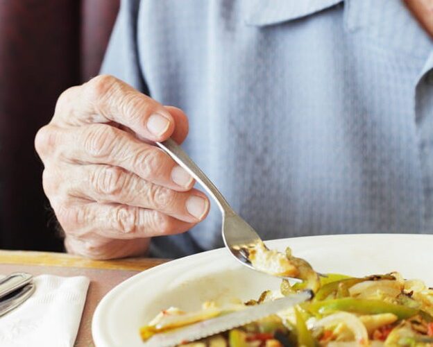 An Upclose Photo Of An Elderly Man Holding A Fork As He Tries To Eat.