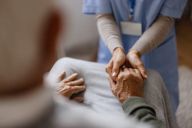 A Nurse Holds The Hand Of An Elderly Man In A Skilled Nursing Facility.